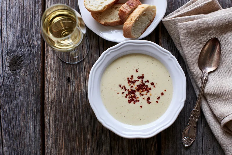 Cream soup with red pepper flakes in a white vintage bowl on a dark wooden table with bread, wine and silver spoon