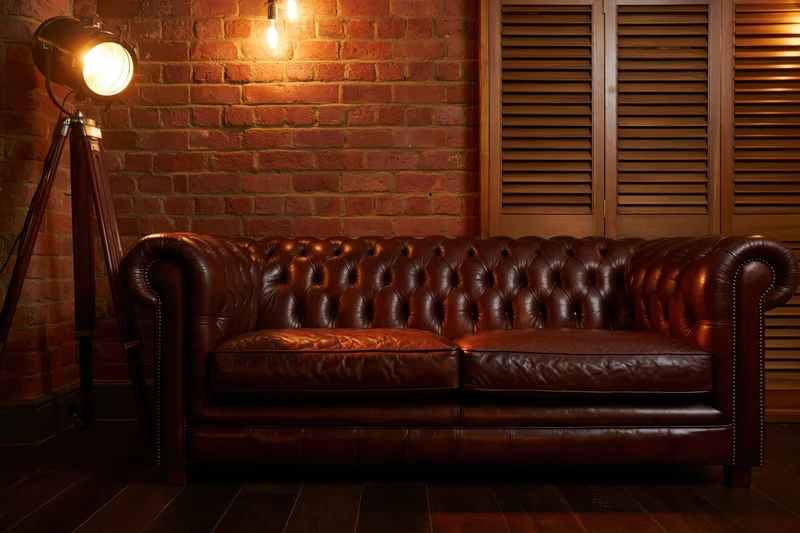 Dark brown tufted chesterfield leather sofa against brick wall with spotlight lighting in rustic living room