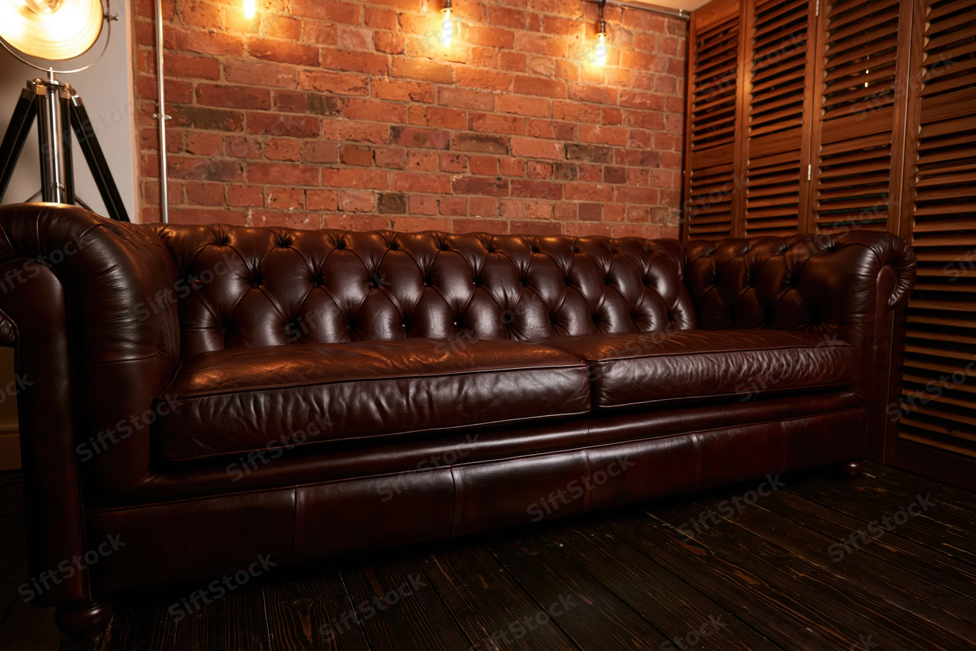 Dark brown tufted chesterfield leather sofa against exposed brick wall in dimly lit room with wooden flooring and warm lighting