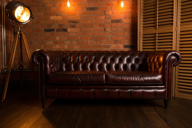 Dark brown tufted chesterfield leather sofa against brick wall with spotlight and wooden floor