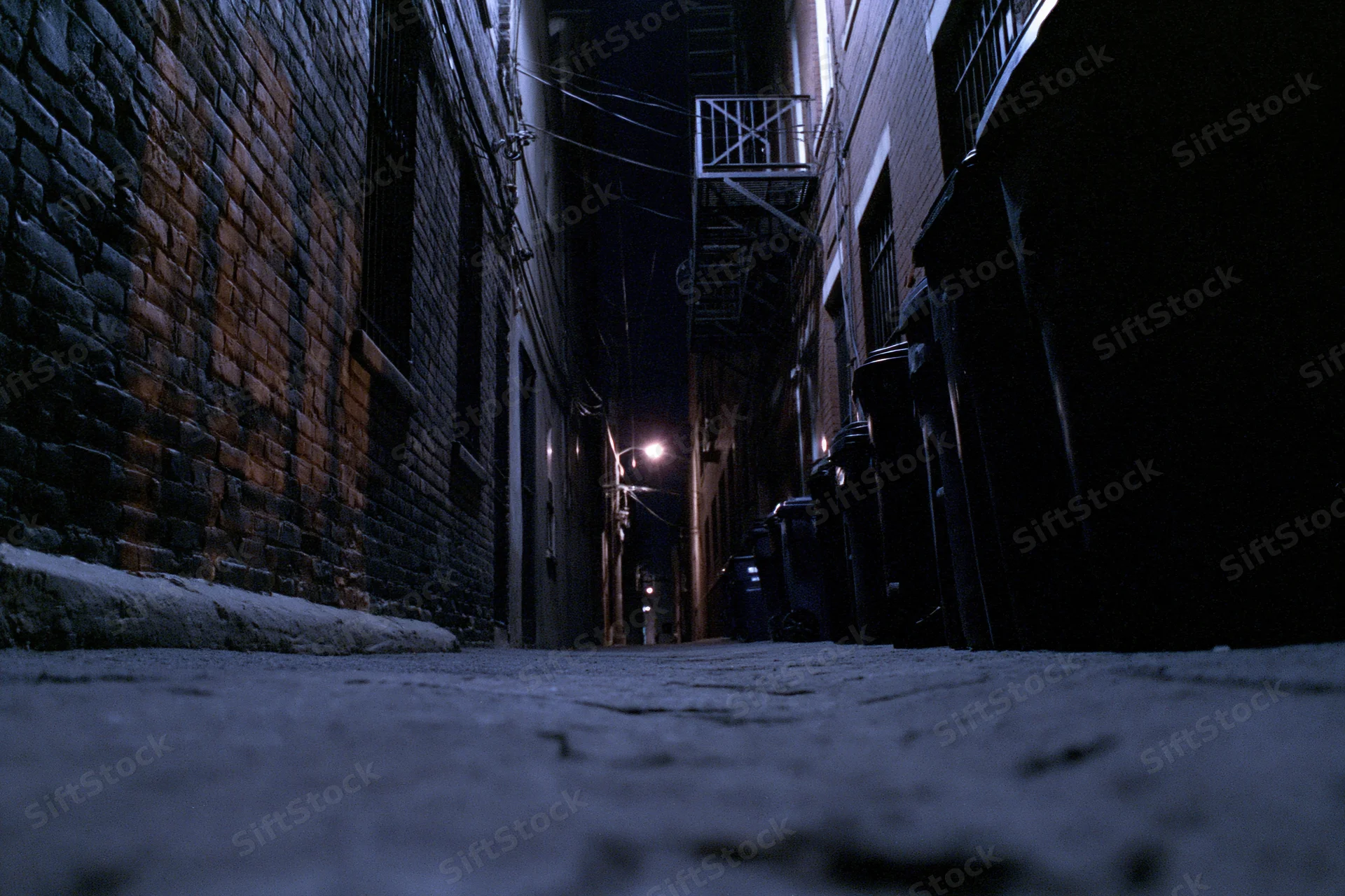 Low-angle view of empty dark alley at night with trash bins lined against brick walls and streetlight in distance
