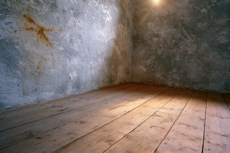 Empty corner of a room with weathered concrete walls and worn wooden floor under dim light