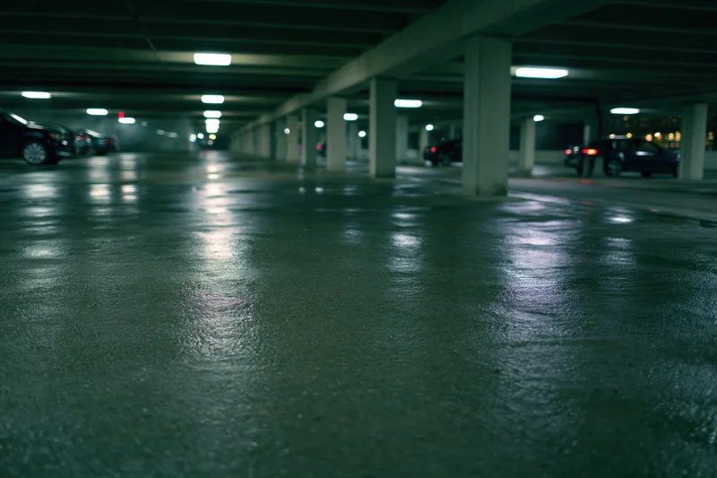 Wet concrete floor in dimly lit underground parking garage with pillars and fluorescent lights