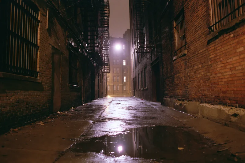 Narrow urban alley at night with wet pavement reflecting distant light