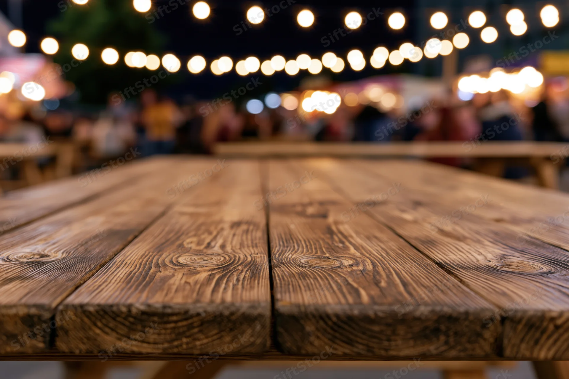 Wooden picnic table surface in sharp focus with blurred lights and people in background at night