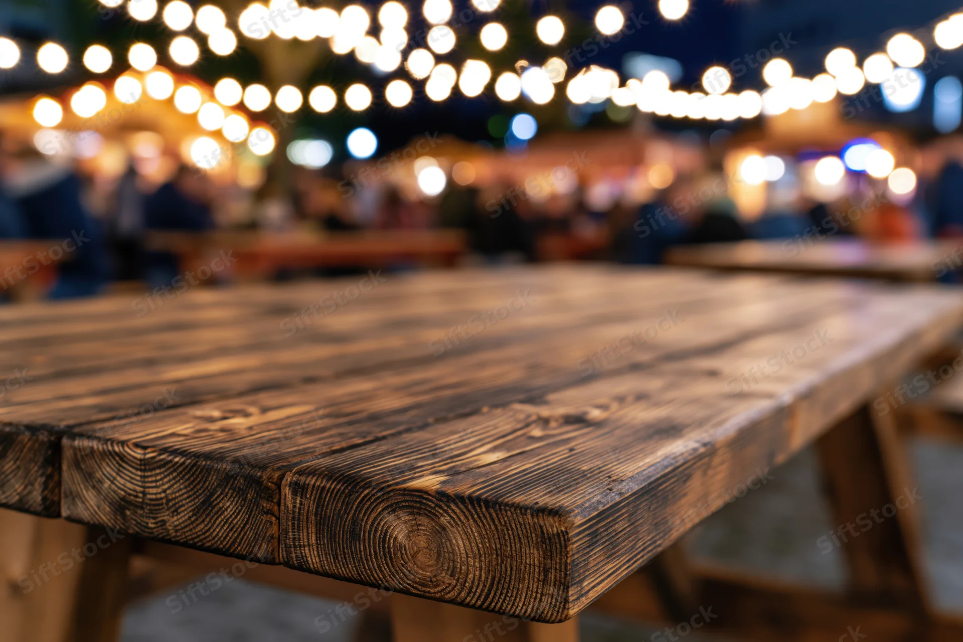 Wooden picnic table surface in sharp focus with blurred string lights and people in background at night