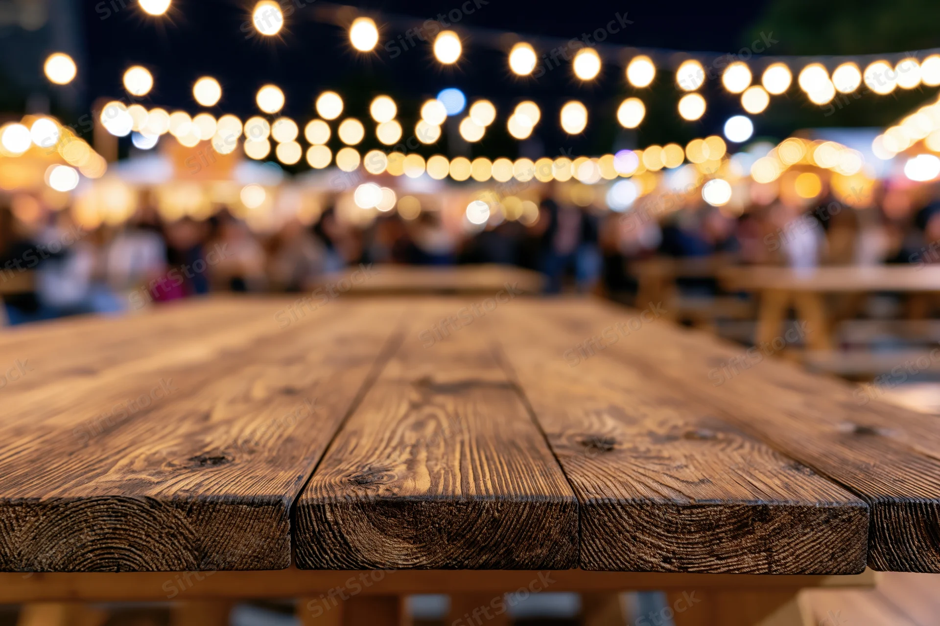 Wooden picnic table surface in sharp focus with blurred string lights and people in background at night