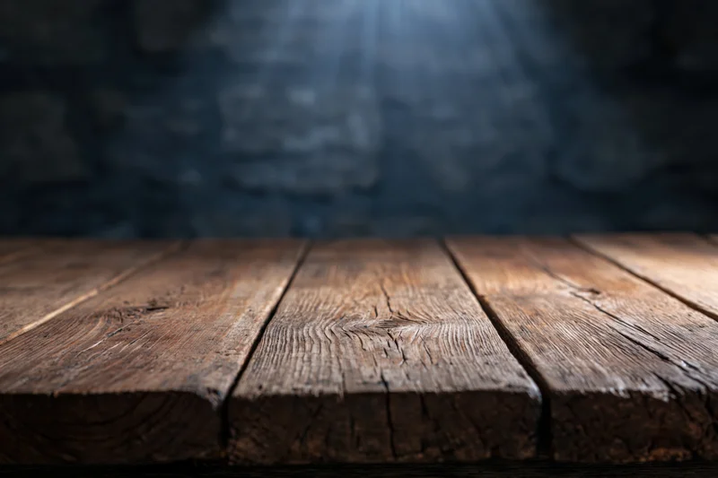 Worn wooden tabletop in a dark room with spotlight and blurred stone wall background