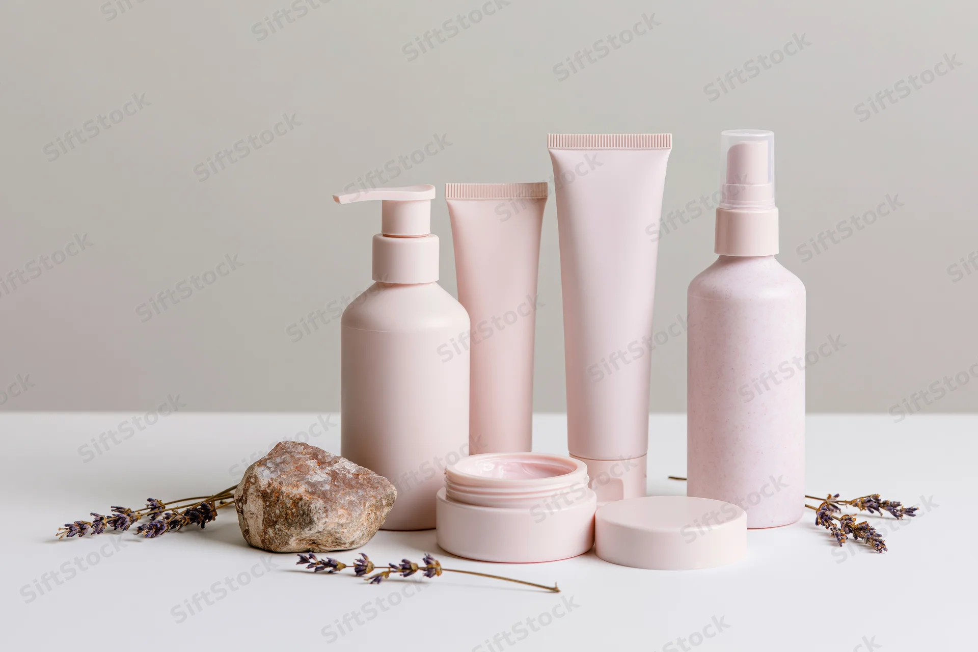 Blush pink skincare containers arranged with lavender and rock on white surface