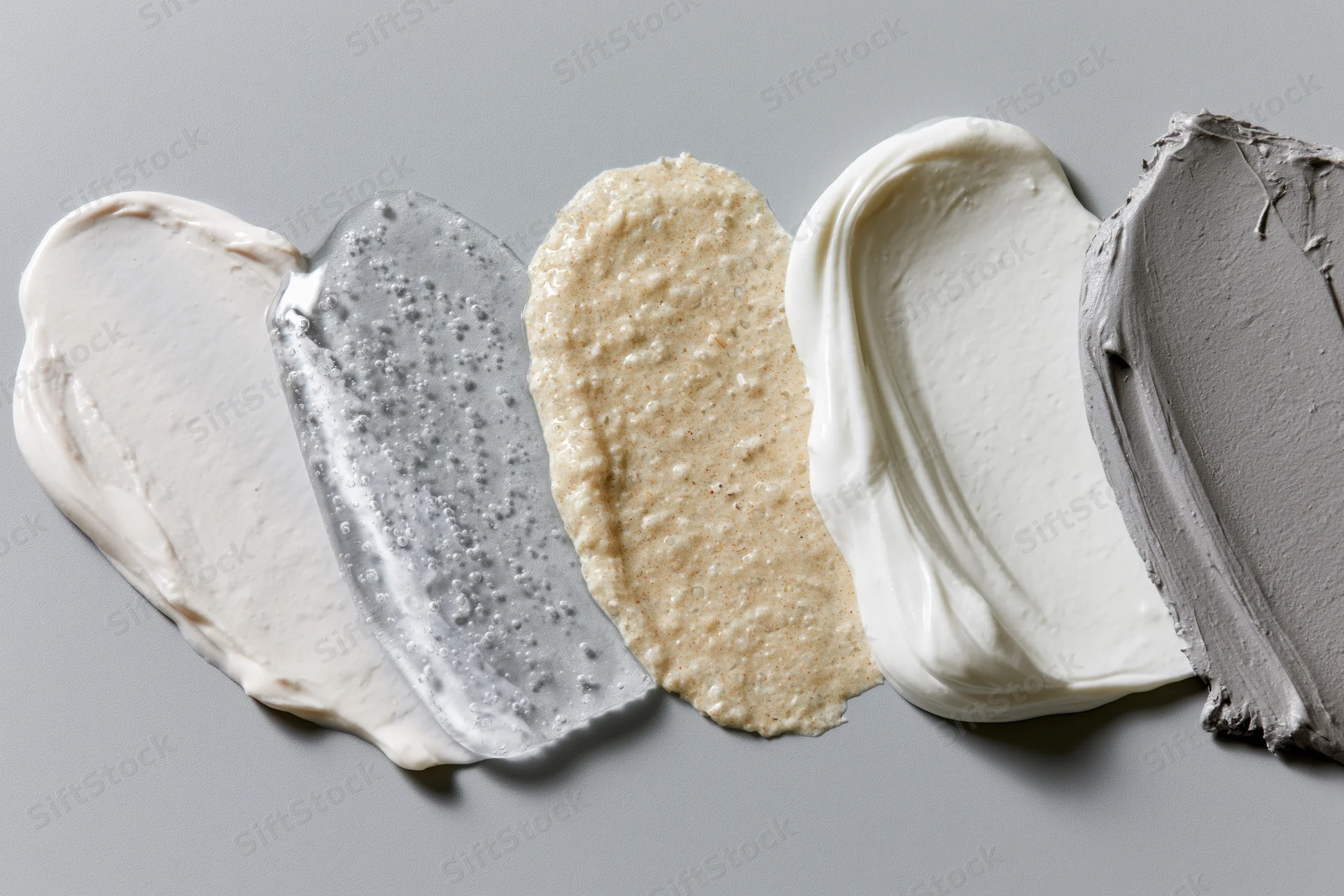Five skincare cream smears arranged in a row on a flat gray surface