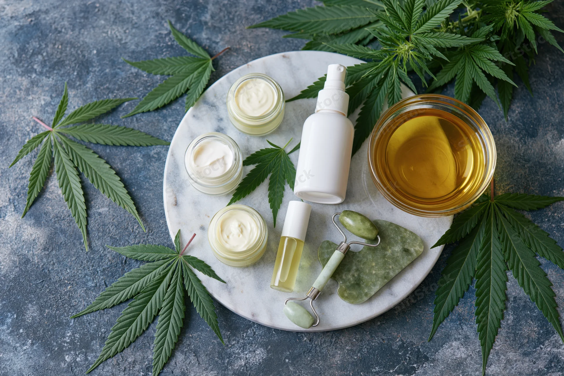Cbd skincare products arranged on a round marble tray with cannabis leaves and jade tools