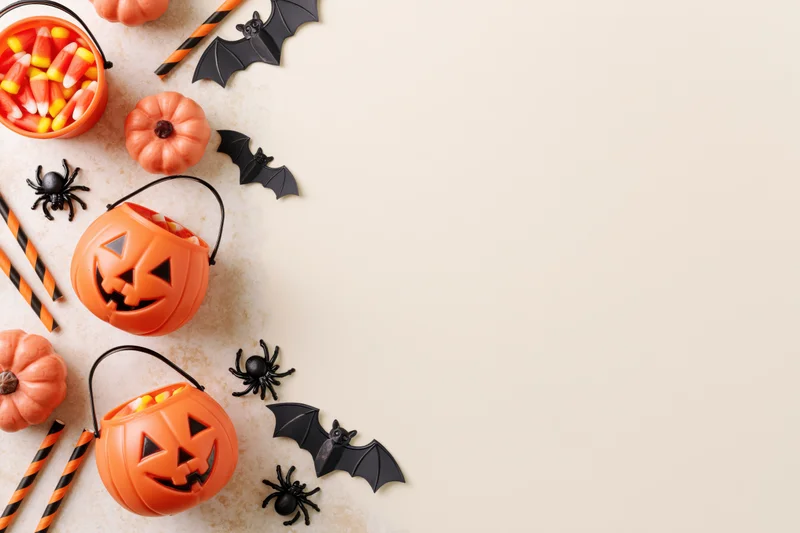 Halloween decorations including pumpkins, bats, spiders, and candy arranged in flat lay on light surface with copyspace on right