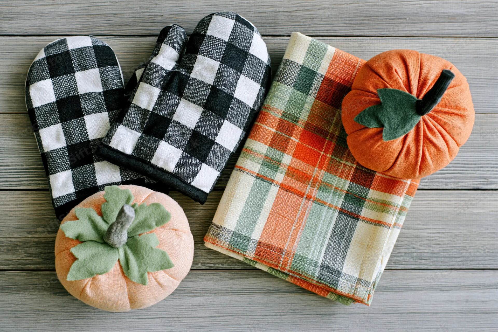 Flat lay of black and white checkered oven mitts, plaid dishcloth, and fabric pumpkins on wooden surface