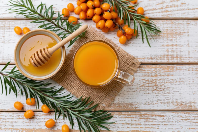 Glass mug of sea buckthorn juice with honey and sprigs on white wooden table