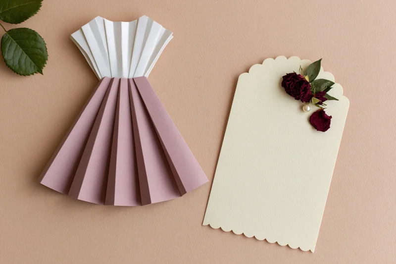 Handmade paper dress craft with scalloped card and dried roses on beige surface