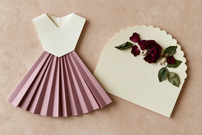 Handmade paper dress craft with scalloped card and dried roses on textured surface