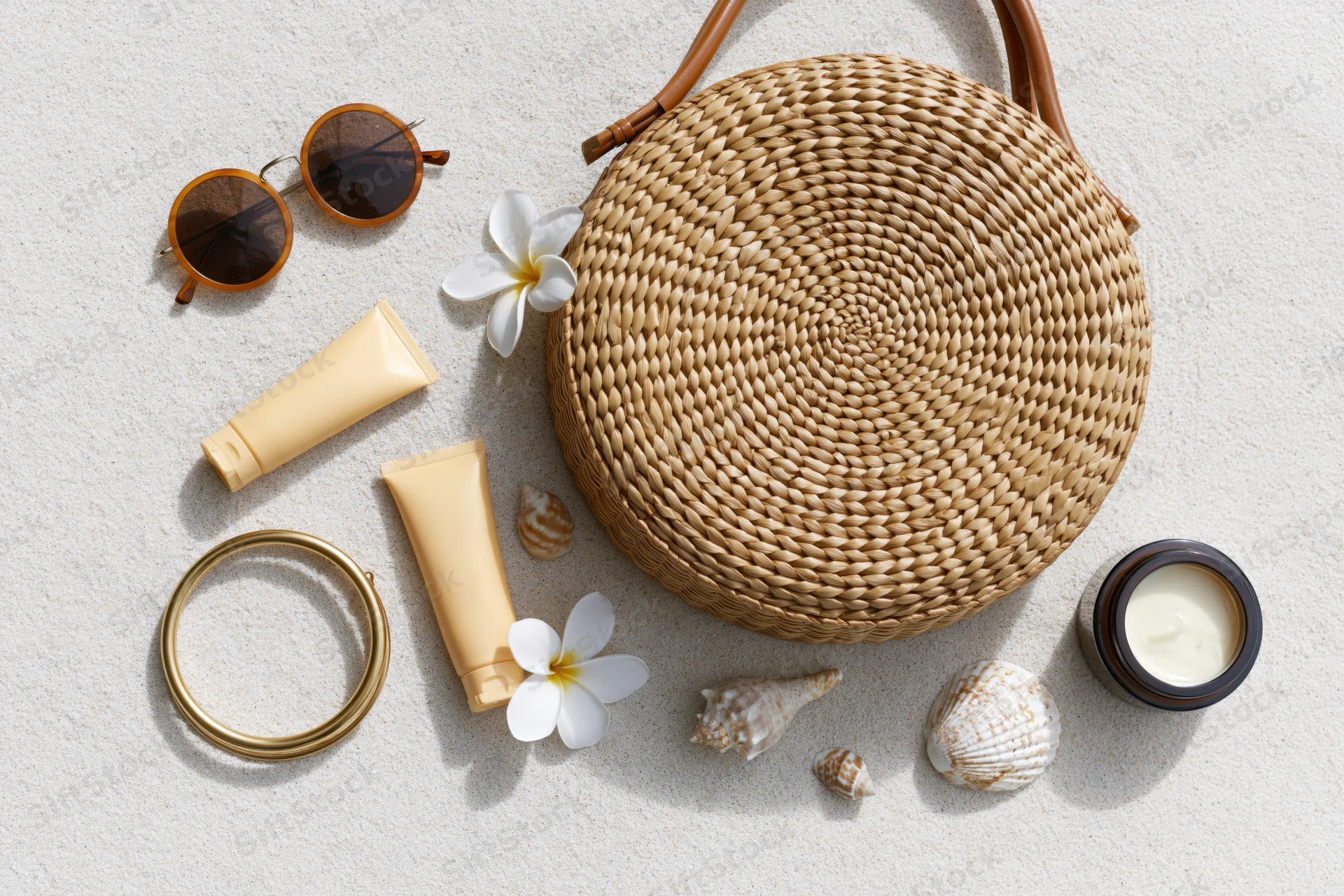Round woven rattan bag with sunglasses, cream tubes, shells and flowers arranged on sand in flat lay style