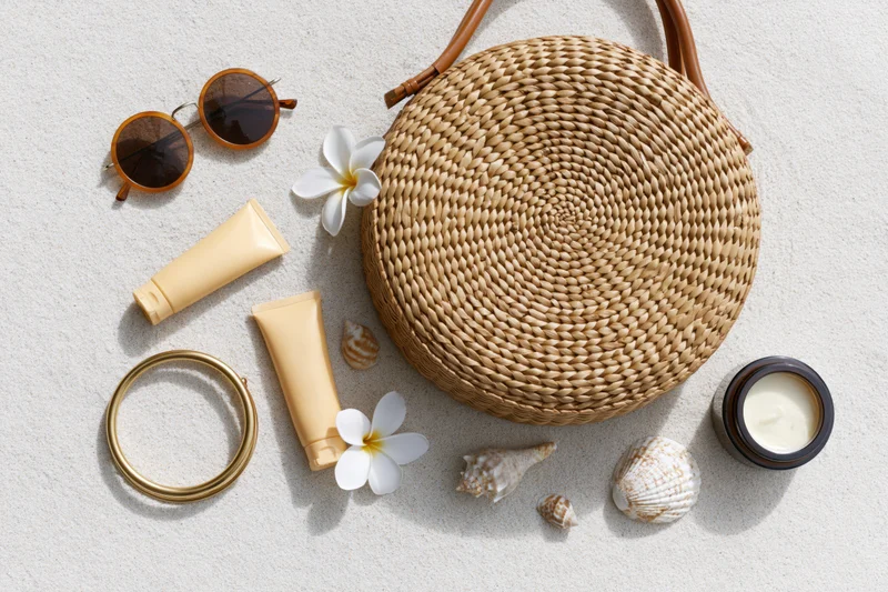 Round woven rattan bag with sunglasses, cream tubes, shells and flowers arranged on sand in flat lay style