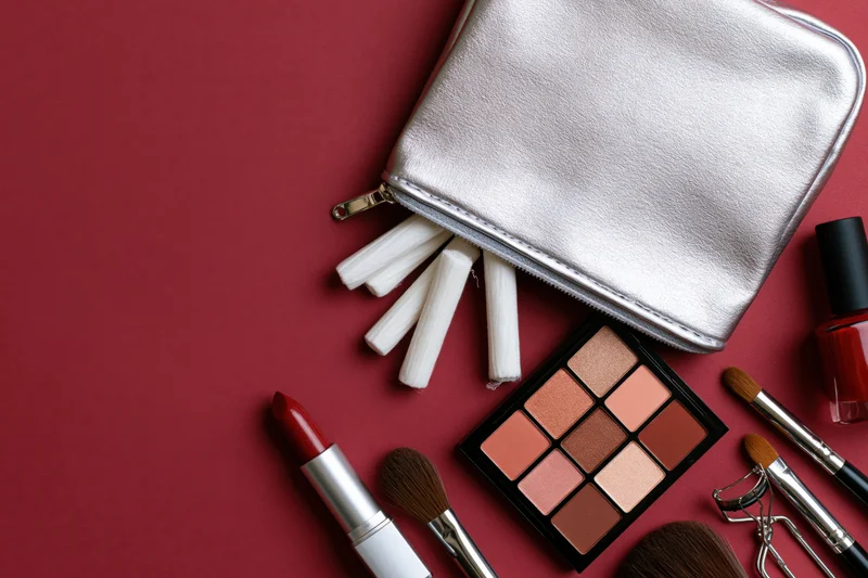 Silver makeup bag with spilled contents including lipstick, eyeshadow palette, brushes and nail polish on red background
