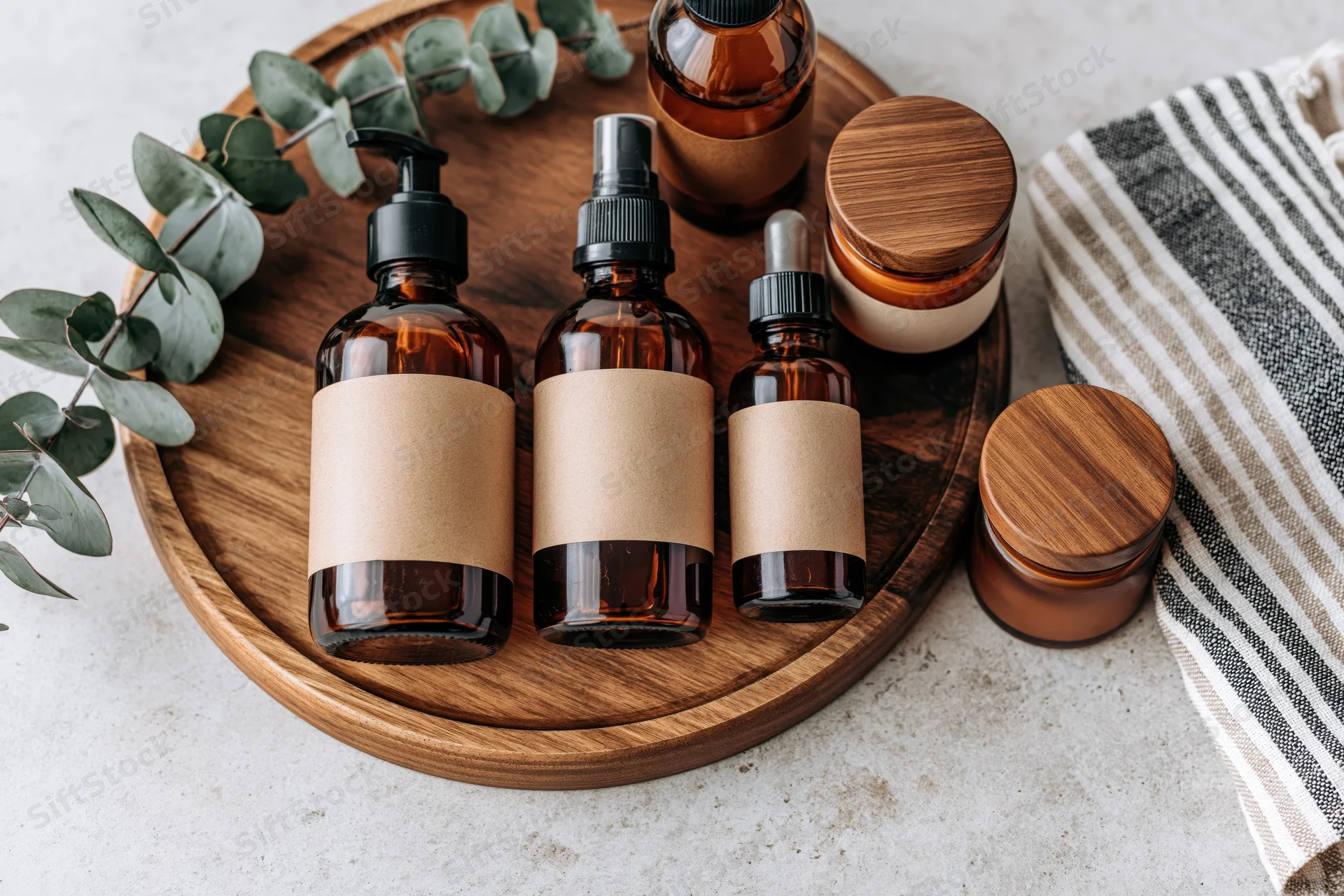 Amber glass skincare bottles with blank kraft labels arranged on a wooden tray with eucalyptus leaves and a striped towel on a marble surface