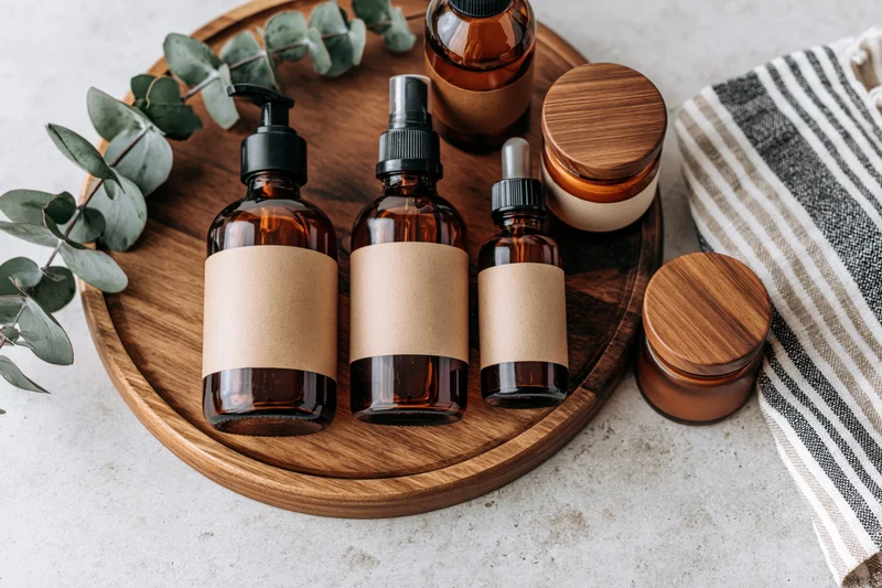 Amber glass skincare bottles with blank kraft labels arranged on a wooden tray with eucalyptus leaves and a striped towel on a marble surface