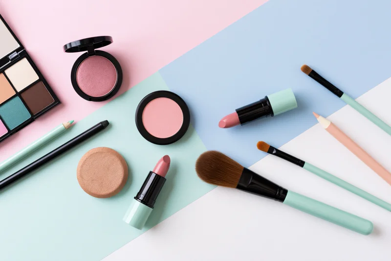 Assorted makeup products arranged in flat lay on pastel colored background