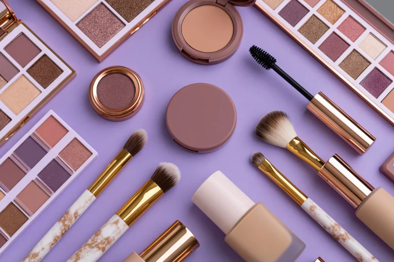 Assorted makeup products including eyeshadow palettes, brushes and compacts arranged in flat lay on purple background