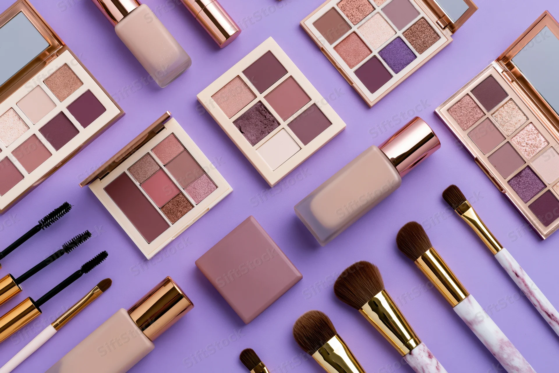 Assorted makeup products including eyeshadow palettes, brushes, foundation and mascara arranged in flat lay on purple background