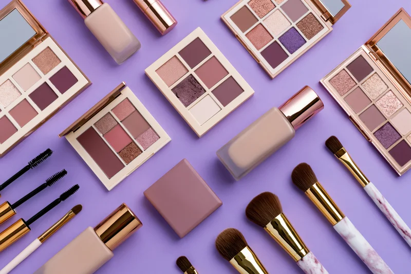 Assorted makeup products including eyeshadow palettes, brushes, foundation and mascara arranged in flat lay on purple background