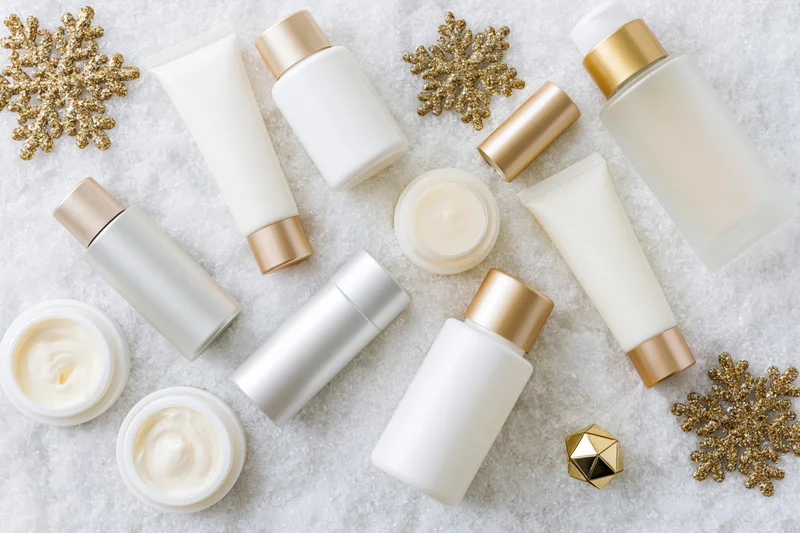Assorted white and silver skincare products arranged on snowy surface with gold snowflake decorations