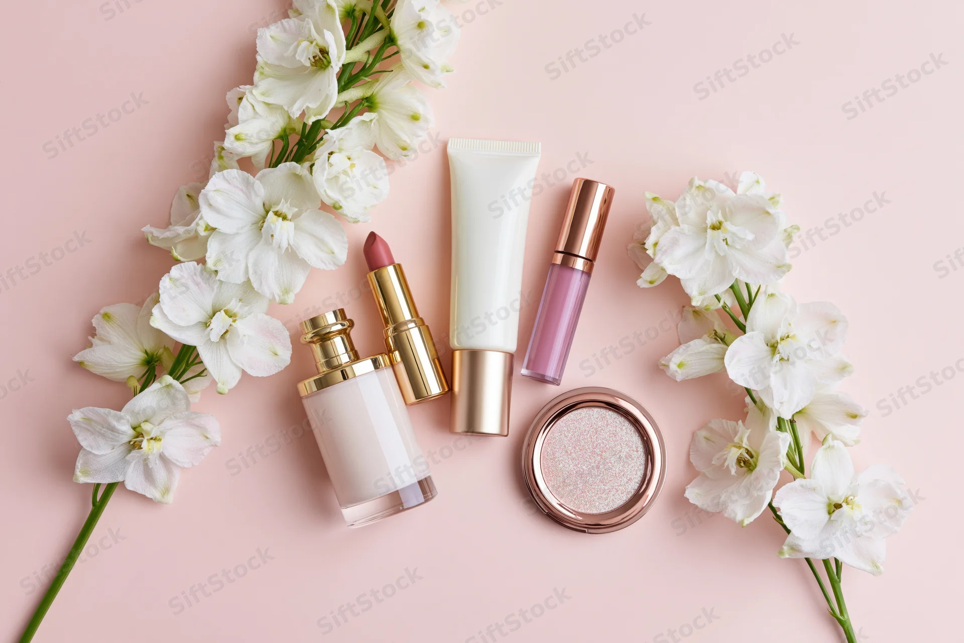 Beauty products and white flowers arranged in a flat lay on a pastel pink surface