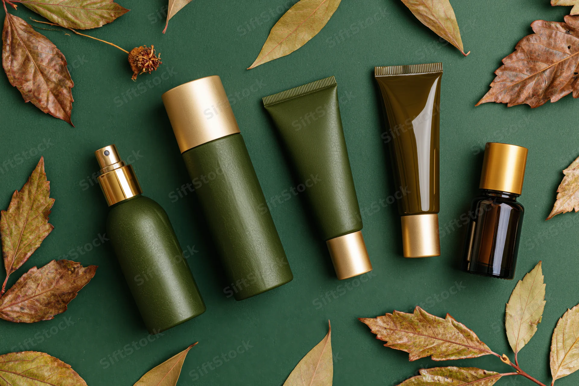 Five green and gold cosmetic bottles and tubes arranged on a dark green surface with dry autumn leaves