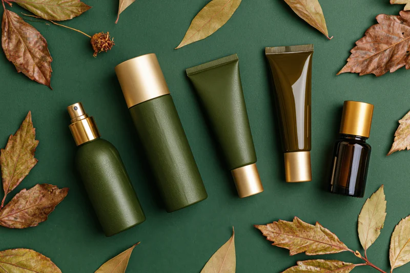 Five green and gold cosmetic bottles and tubes arranged on a dark green surface with dry autumn leaves