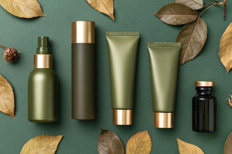 Five unlabeled green and dark olive cosmetic containers arranged in a flat lay with dried leaves on a dark green background