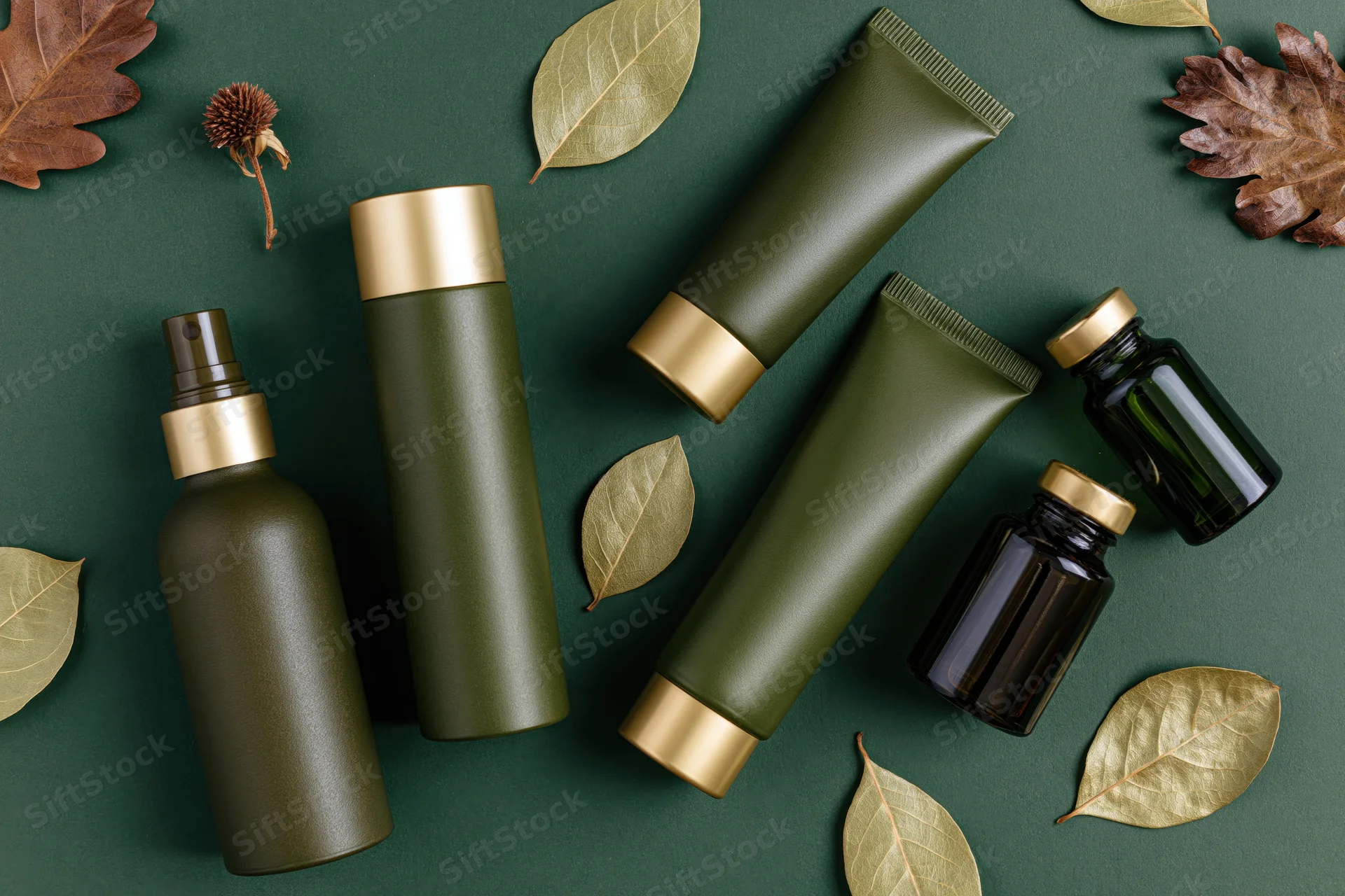 Green and dark olive cosmetic bottles and tubes arranged with dried leaves on a dark green surface in a flat lay top view