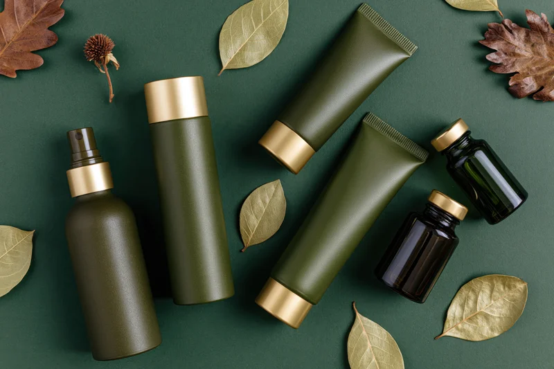 Green and dark olive cosmetic bottles and tubes arranged with dried leaves on a dark green surface in a flat lay top view
