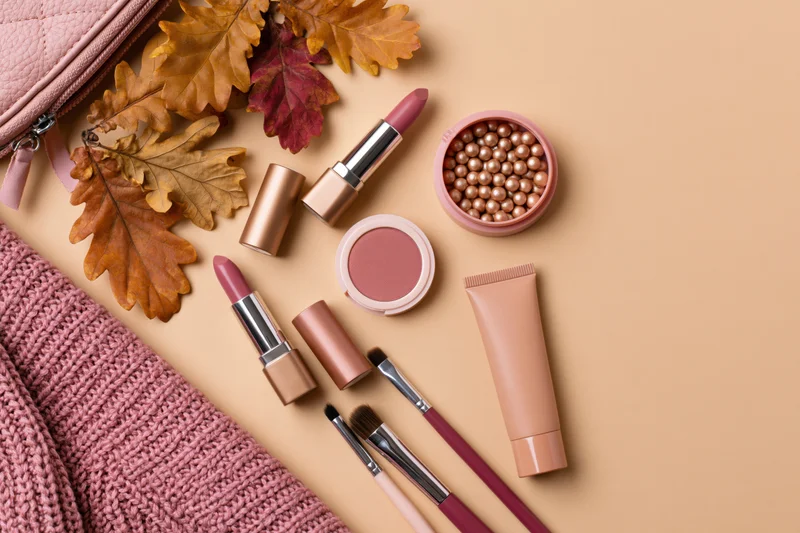 Makeup products and autumn leaves arranged on a beige surface with a knitted sweater and bag