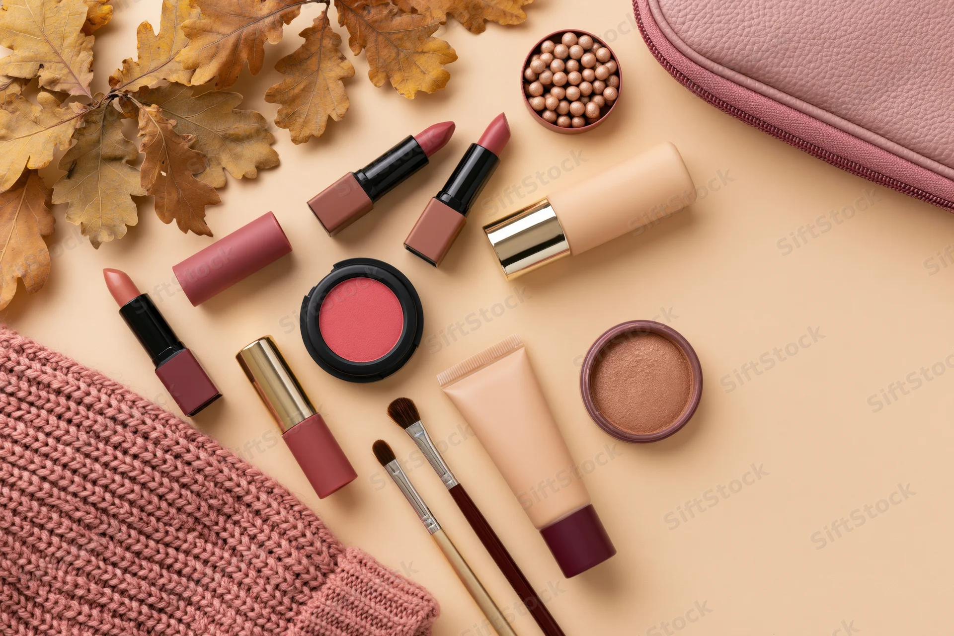 Makeup products arranged with autumn leaves and knit fabric on a beige surface