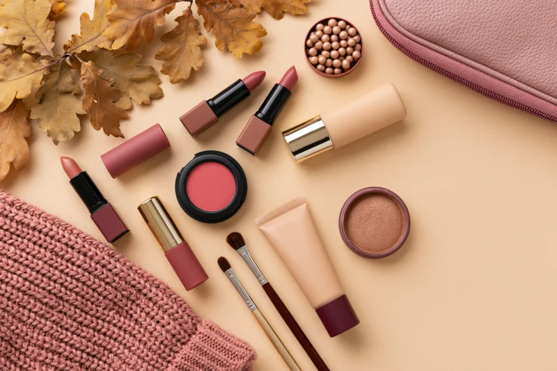 Makeup products arranged with autumn leaves and knit fabric on a beige surface
