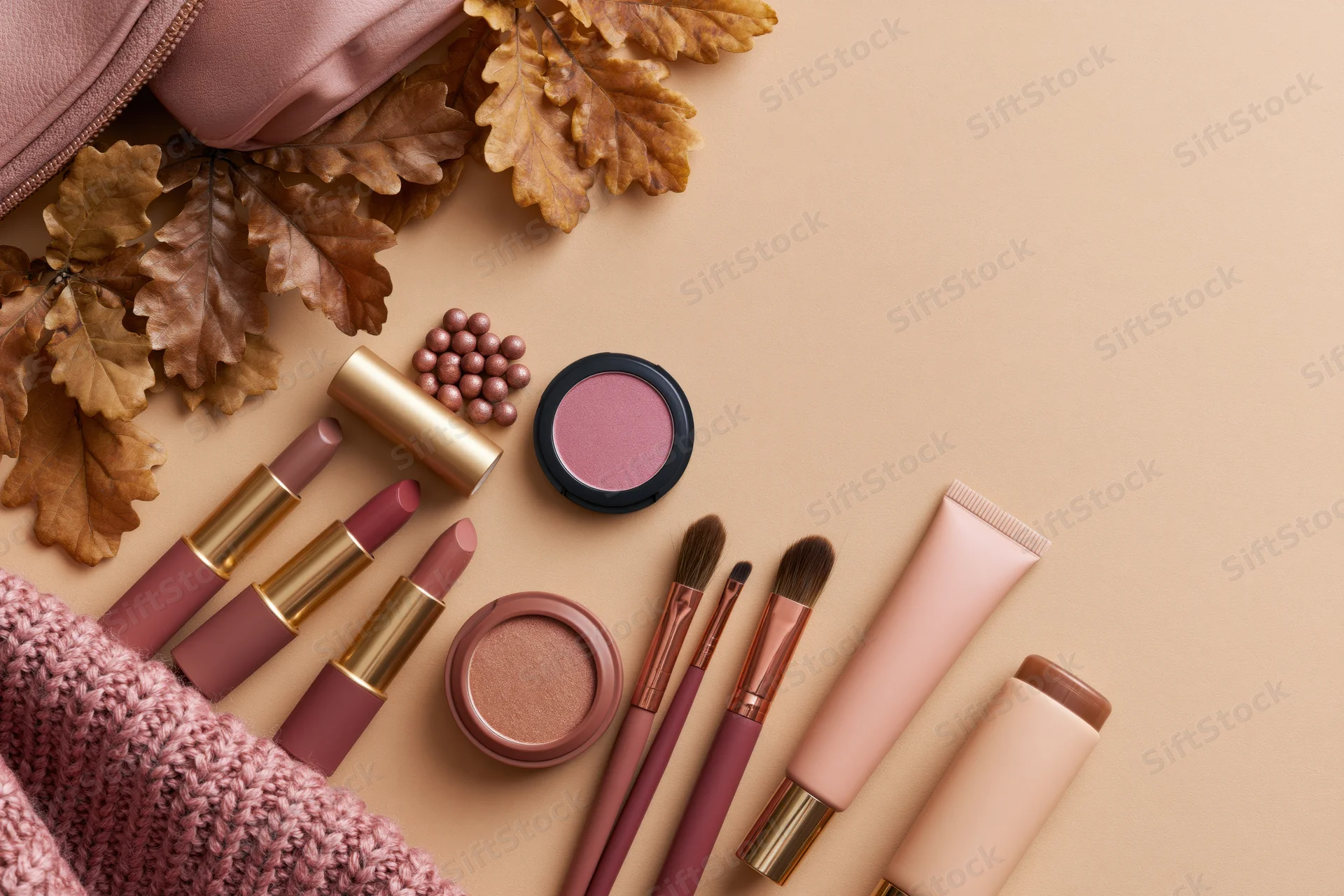 Makeup products arranged with autumn leaves and a knitted bag on a beige surface