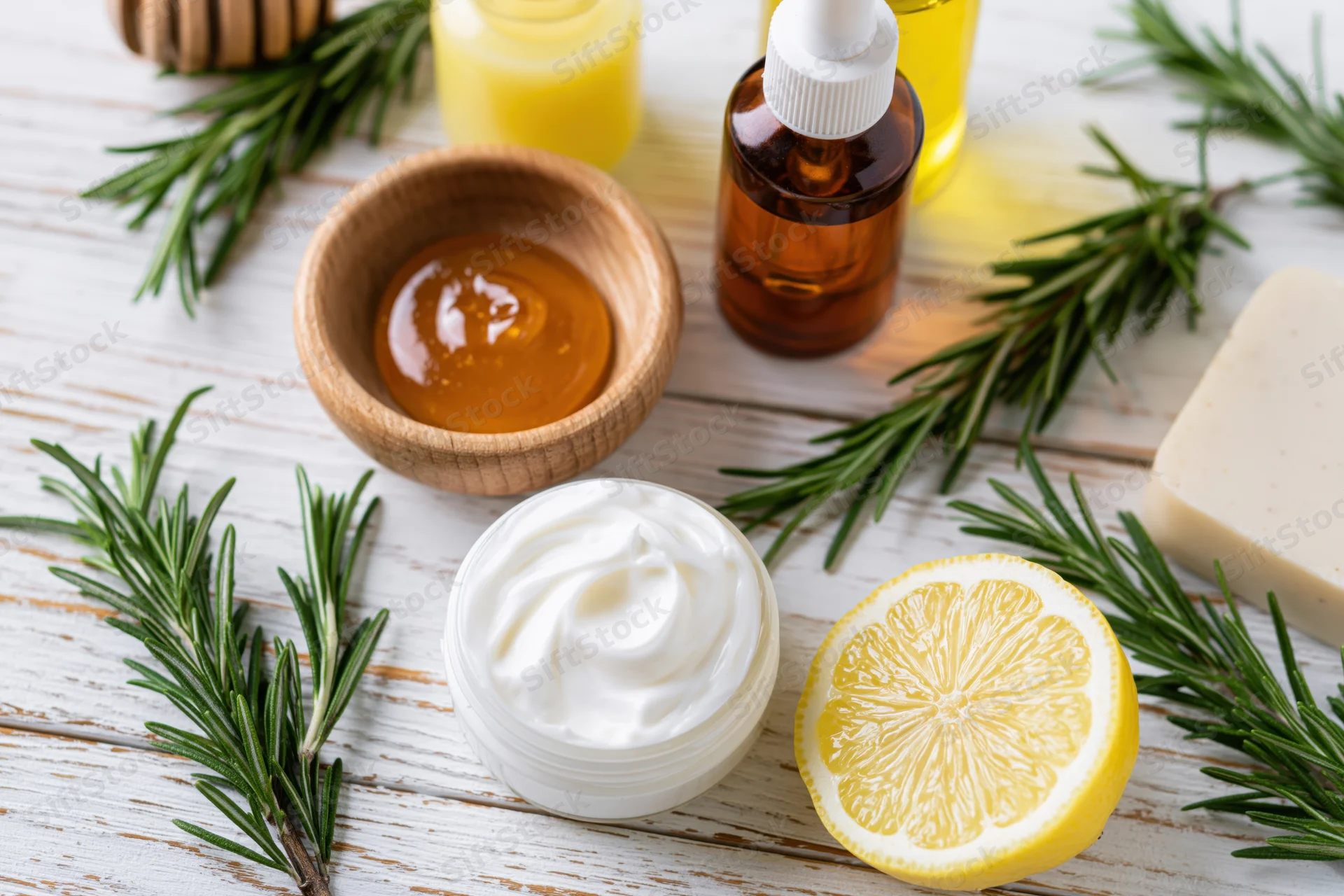 Natural skincare ingredients including rosemary, lemon, honey, cream, and soap arranged in flat lay on white wooden table