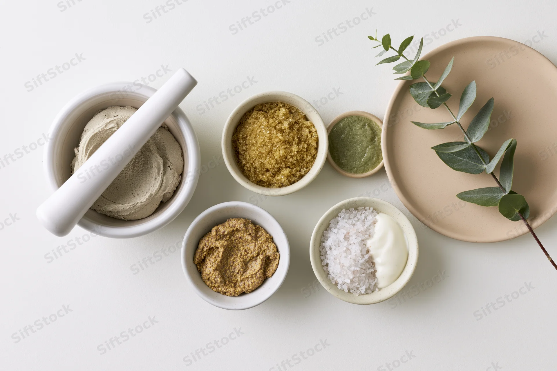 Natural skincare ingredients arranged in ceramic bowls and a mortar with eucalyptus on white surface