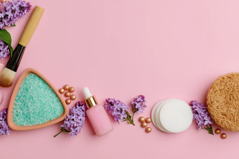Skincare and beauty products arranged in a flat lay on a pink background with lilac flowers and copyspace