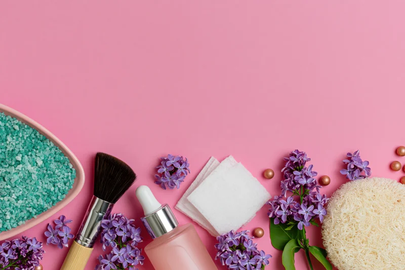 Skincare and beauty products arranged along the bottom of a pink background with lilac flowers and copyspace above