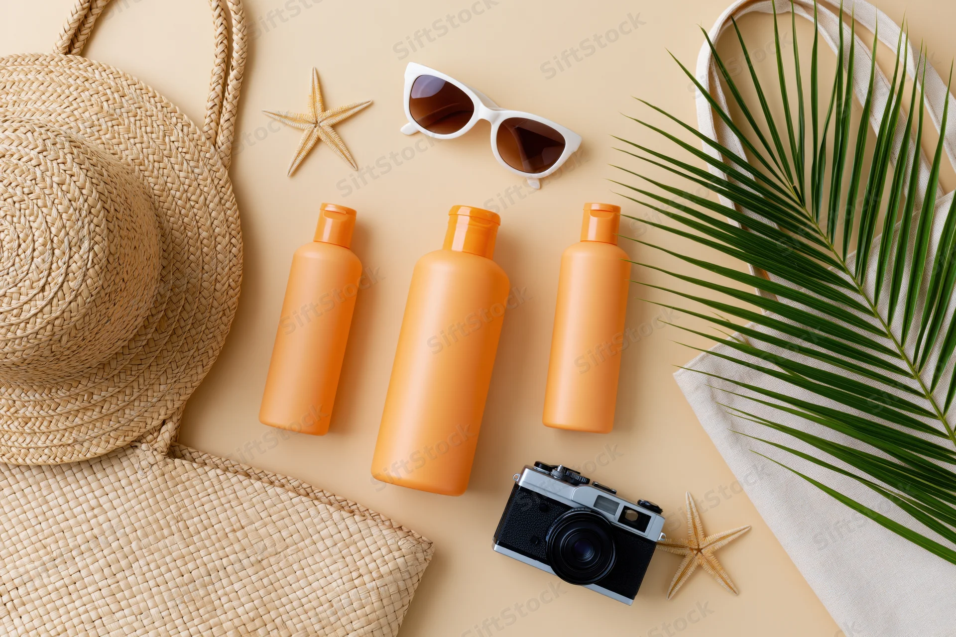 Summer beach accessories arranged flat on beige surface with straw hat, sunglasses, sunscreen bottles, camera, palm leaf and starfish
