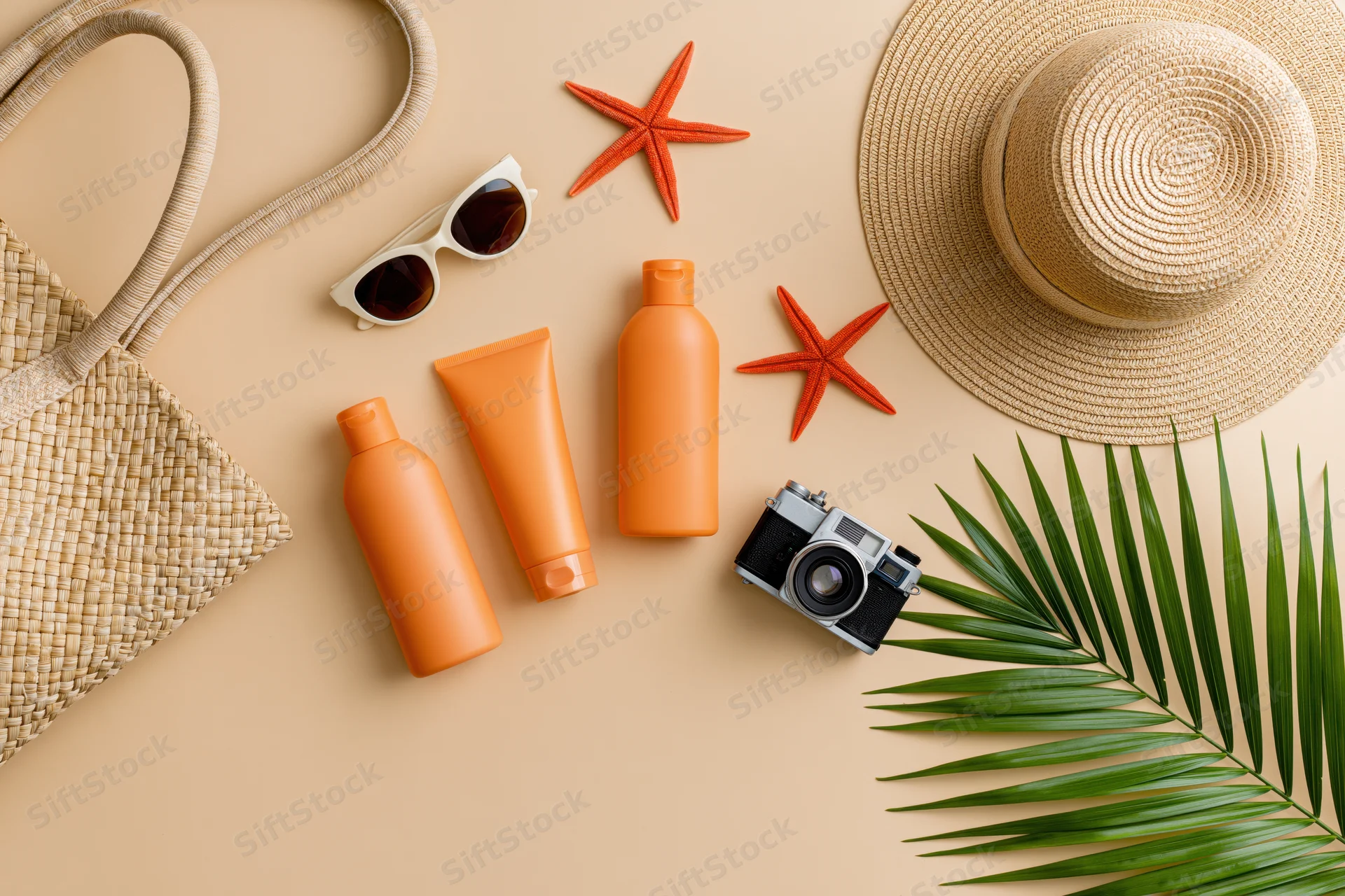 Summer beach accessories arranged flat on warm beige surface with straw hat, sunglasses, sunscreen bottles, camera, palm leaf and starfish