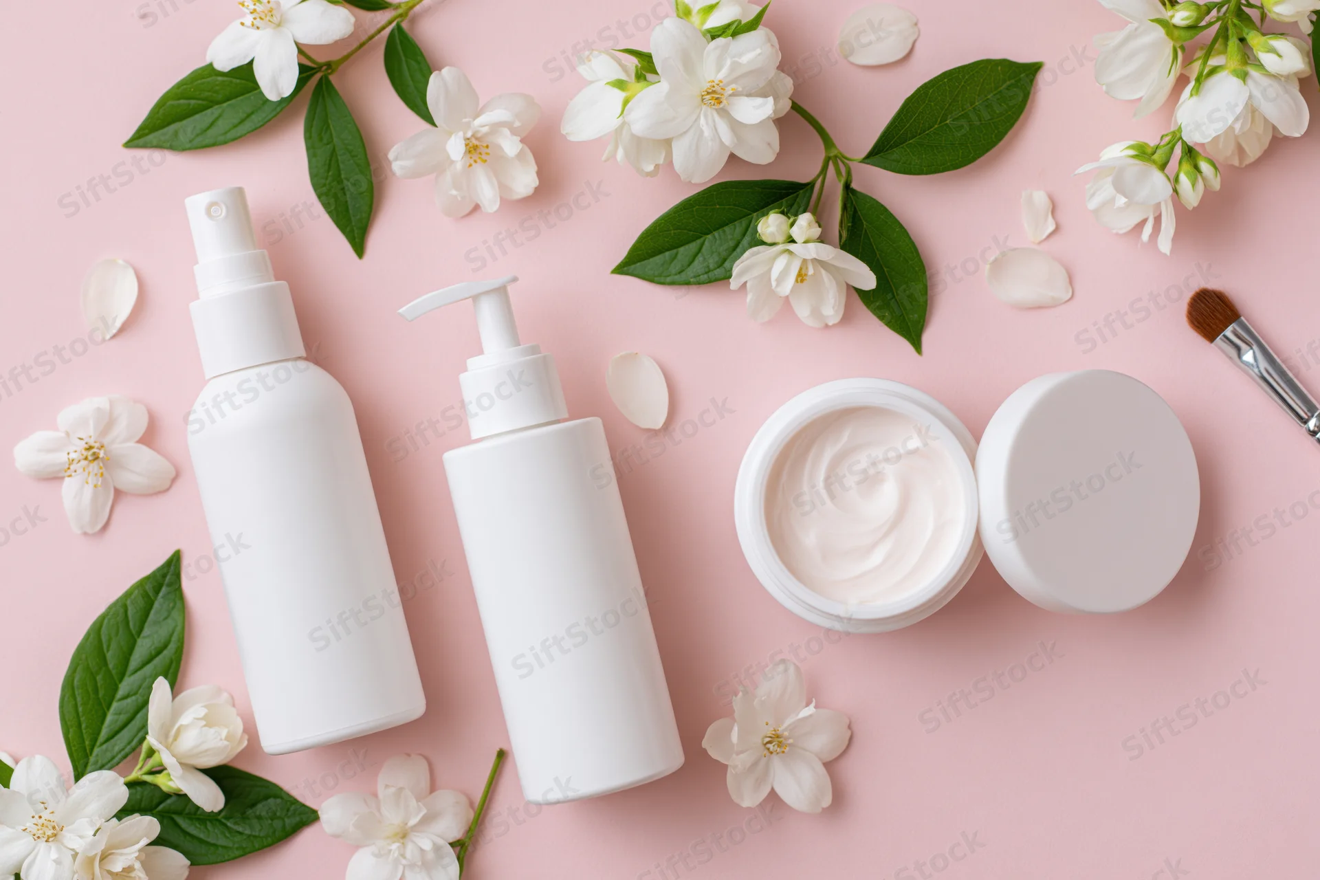 Three white skincare containers with jasmine flowers and a brush on a pink background