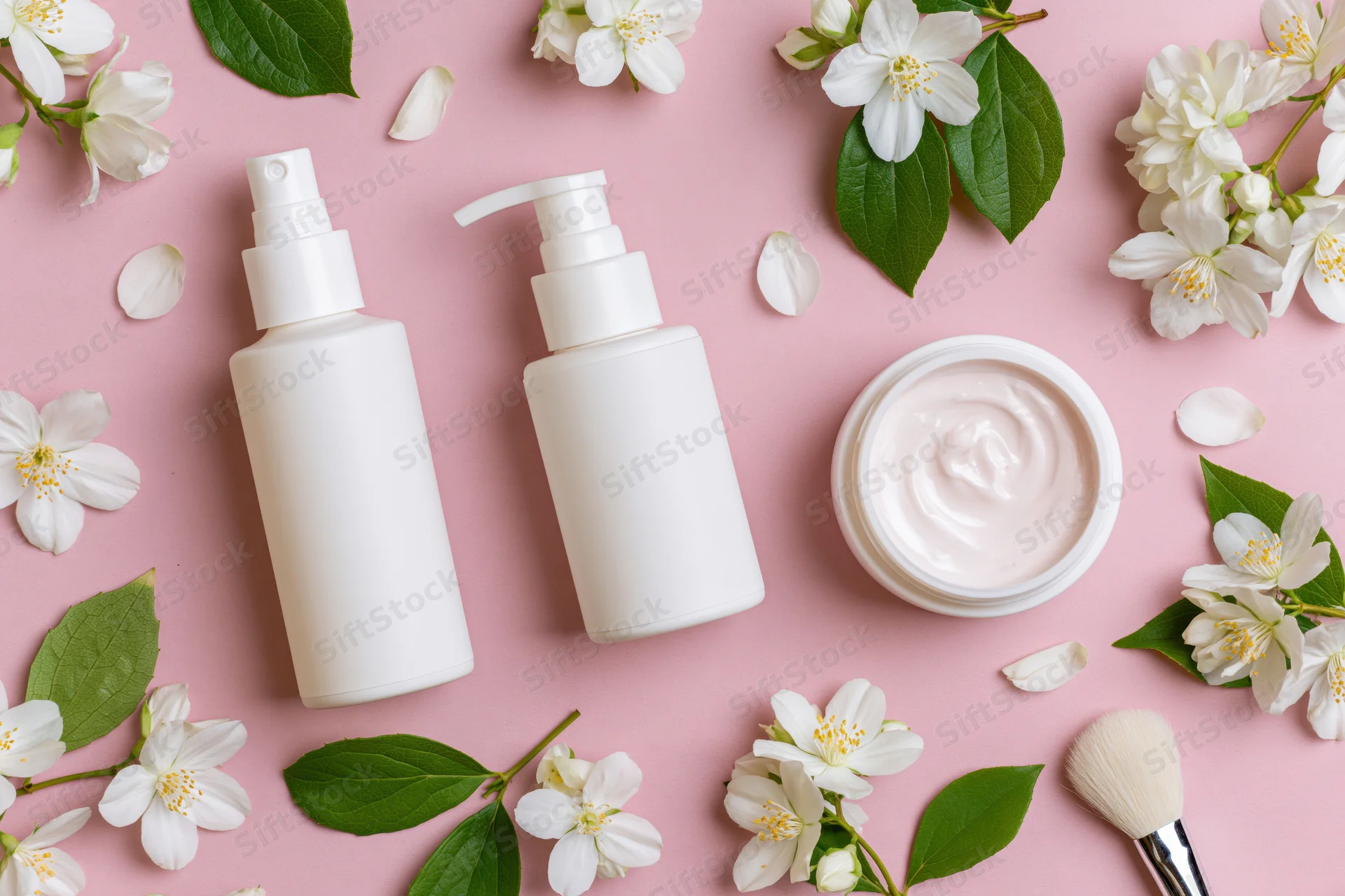 Three white unlabeled skincare containers with jasmine flowers and a brush on pink background