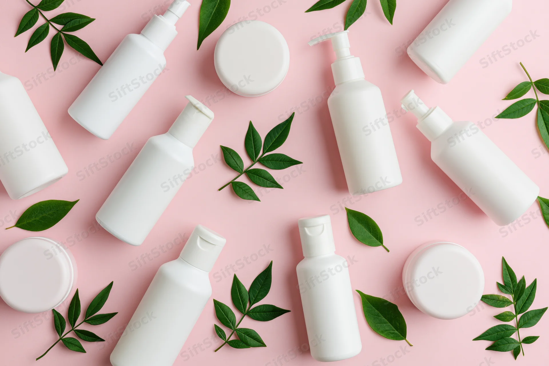 White skincare bottles and green leaves arranged on pink background in flat lay composition