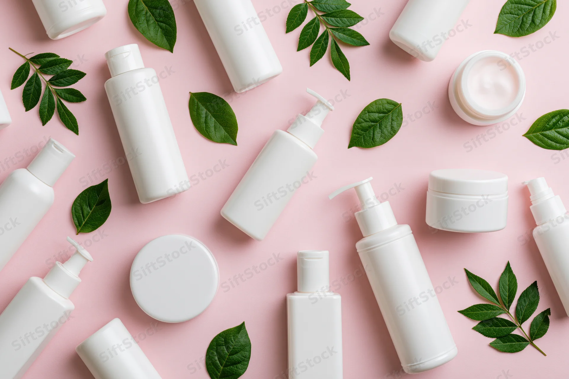White skincare bottles and jars with green leaves arranged on pink background in flat lay style